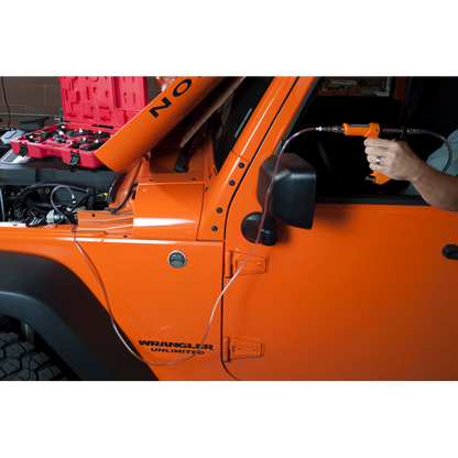 The image shows a man using a tool to bleed the brake fluid of an orange Jeep Wrangler. The brake fluid bleeding gun is connected to a hose that is attached to the brake caliper of the Jeep