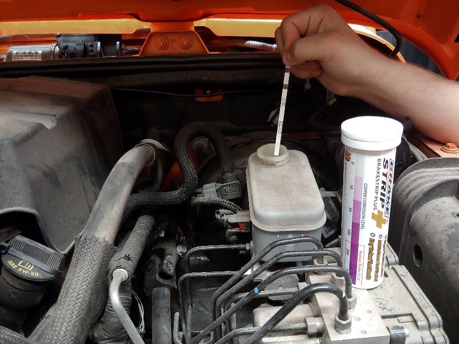 Close-up of car brake fluid reservoir with a dipstick labeled "BrakeStrip Plus" measuring the fluid level. It has a scale from 0 (low) to 100 (full). Text on the reservoir warns about copper corrosion and brake fluid replacement.