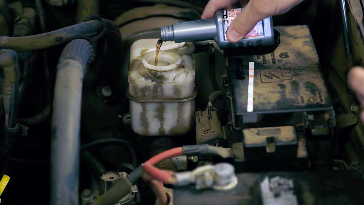 The image shows a person pouring liquid from a BrakeShot Brake Fluid Crash Avoidance Treatment into a car part. The car part has a cylindrical reservoir with a visible minimum fill level line. Brake fluid is a type of hydraulic fluid that transmits pressure from the brake pedal to the car's braking system.