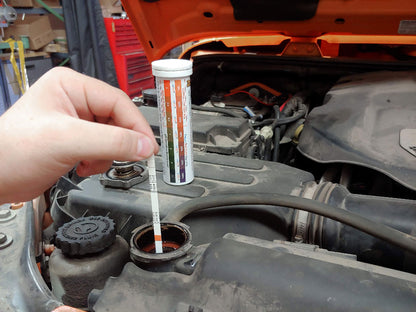 The image depicts a person holding a package of coolant and brake fluid test strips in front of a car engine