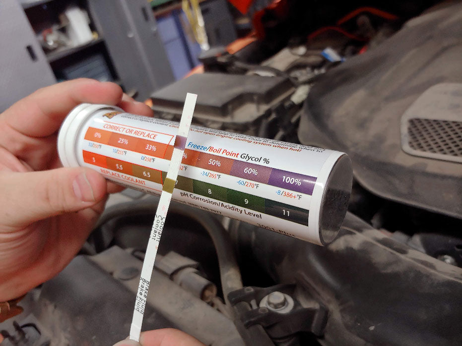 The image depicts a person holding a package of coolant and brake fluid test strips in front of a car engine. Text on the package displays a chart measuring freeze and boiling points,  a glycol percentage scale, and a pH corrosion level.